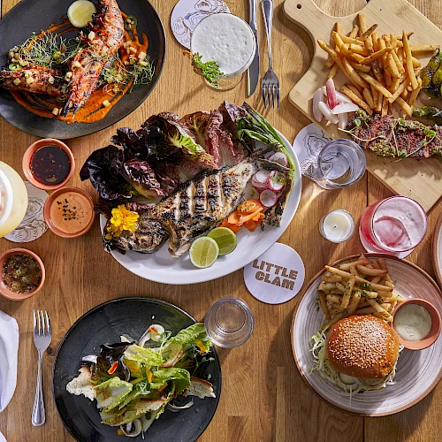 Sizzling plates of steak, burgers, fries, and fresh greens on a rustic wooden table&mdash;delicious spread for sharing.