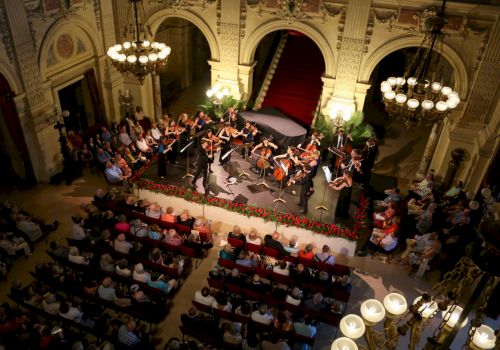 A large orchestra performs on a stage in a grand hall, with a conductor leading, surrounded by an audience in a historic, illuminated venue.