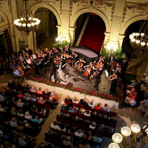 A large orchestra performs on a stage in a grand hall, with a conductor leading, surrounded by an audience in a historic, illuminated venue.