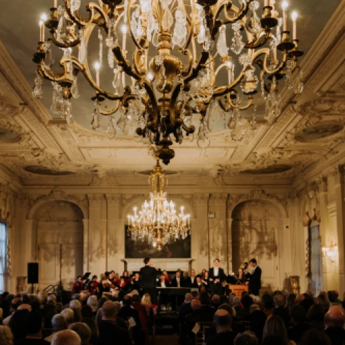 A grand concert hall with ornate ceilings and chandeliers, a pianist on stage, and a large audience seated for a formal performance.