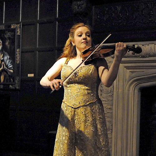 A woman in a glittery gold gown plays a violin on a dark stage, spotlighted, with a painted wall and fireplace backdrop, focused and elegant.