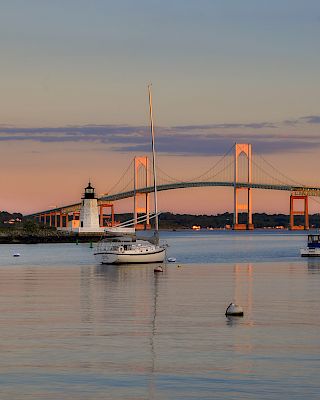 A calm harbor at dusk with several sailboats anchored near the shore and a lit bridge reflecting on the water, pastel sky above.