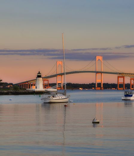 A calm harbor at dusk with several sailboats anchored near the shore and a lit bridge reflecting on the water, pastel sky above.