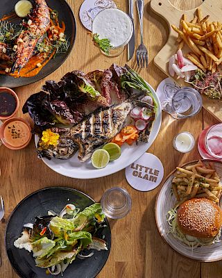 A spread of grilled meats, burgers, salads, fries, and sauces on a wooden table, with drinks and various dipping sauces.
