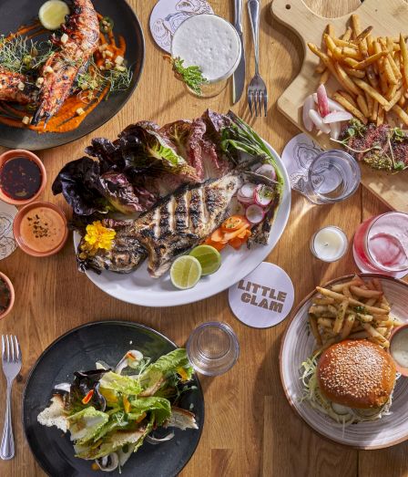 A spread of grilled meats, burgers, salads, fries, and sauces on a wooden table, with drinks and various dipping sauces.