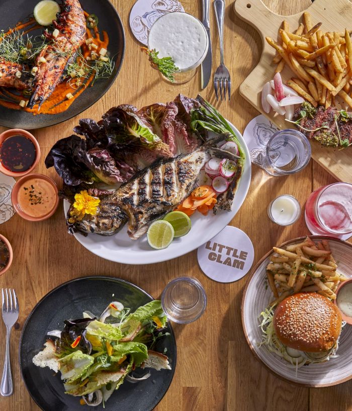 A spread of grilled meats, burgers, salads, fries, and sauces on a wooden table, with drinks and various dipping sauces.