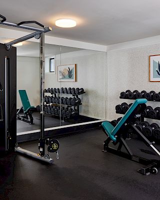 A small gym with a cable machine, a teal adjustable bench, free weights on racks, and a mirrored wall reflecting equipment, plus framed art on the wall.