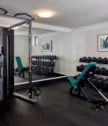 A small gym with a cable machine, a teal adjustable bench, free weights on racks, and a mirrored wall reflecting equipment, plus framed art on the wall.