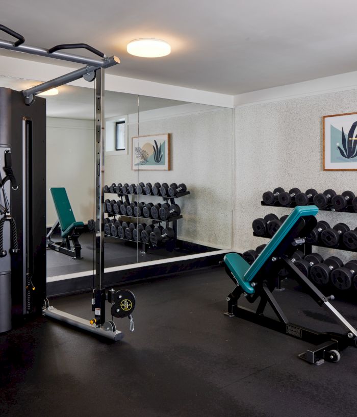 A small gym with a cable machine, a teal adjustable bench, free weights on racks, and a mirrored wall reflecting equipment, plus framed art on the wall.