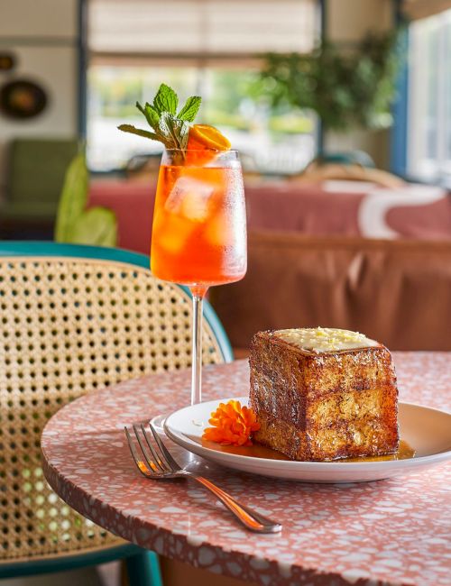 A cafe scene with a pink mosaic table, a slice of cake and orange drink garnished with mint on the plate, in a cozy retro setting, ending with a period.