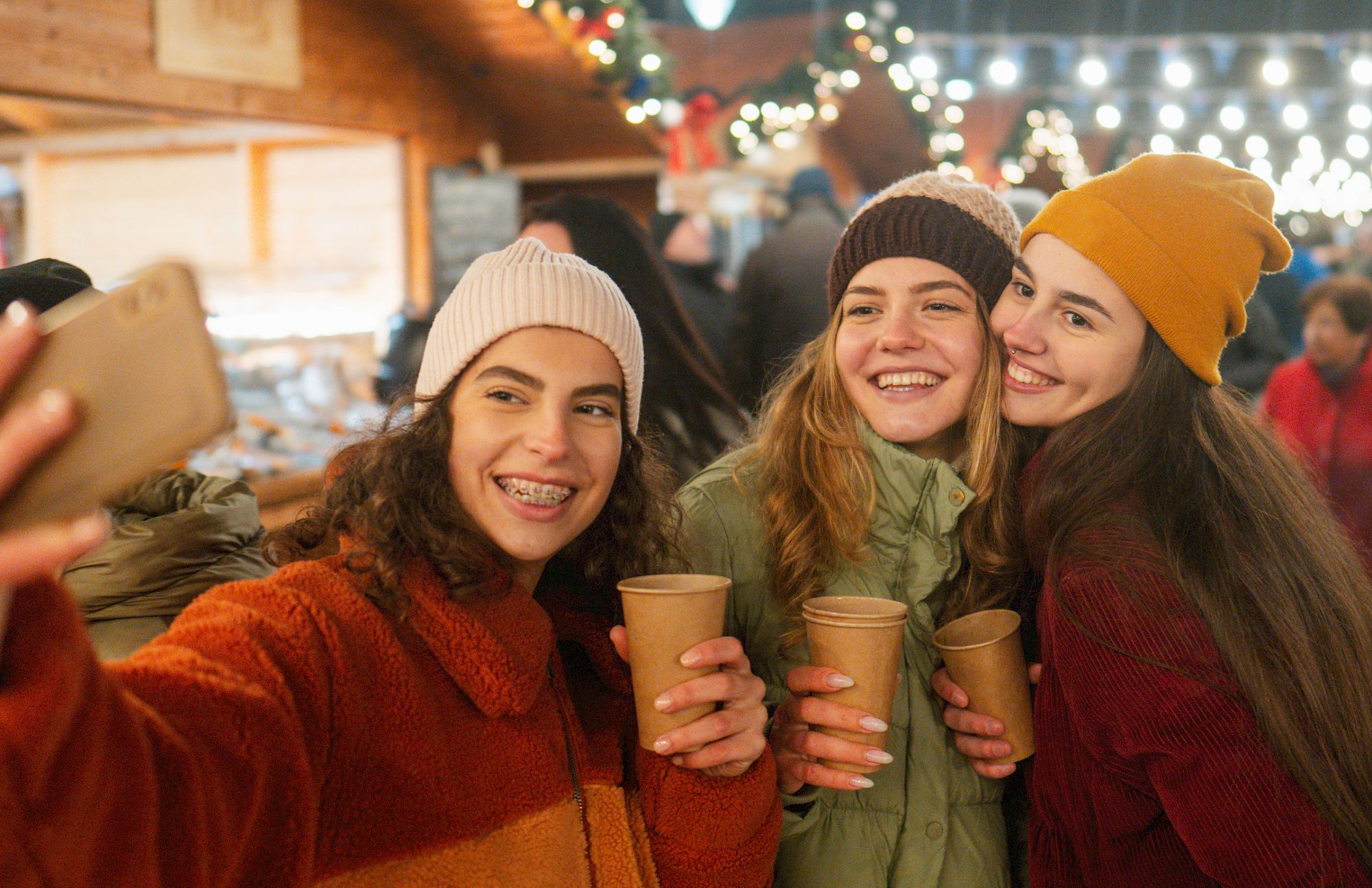 Three friends smiling for a selfie at a festive outdoor market, holding warm drinks and wearing cozy hats under string lights.
