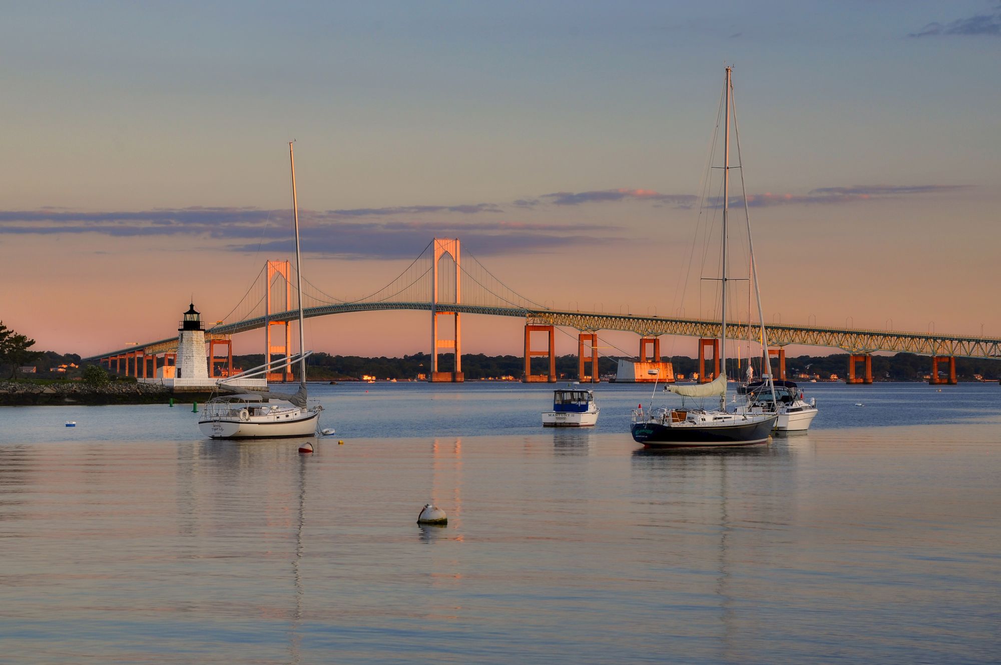 Sailboats float on calm water at sunset with a bridge and lighthouse in the background, reflecting warm orange hues across the scene.