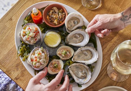 A platter of assorted appetizers and oysters on a wooden table, with two cocktails and hands reaching for bites, top it at 140 characters, always ending the sentence.