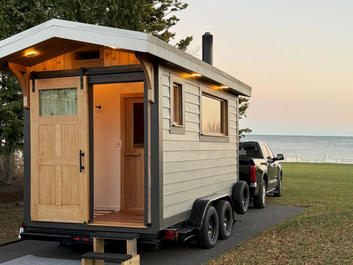 A small, light-gray wooden tiny house on a trailer, parked near a grassy area by the water, with a wooden door open and warm interior light.